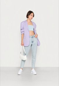 Mulher vestindo um blazer lilás claro, um top de tubo azul, jeans azul claro, tênis brancos, segurando uma pequena bolsa branca, de pé contra um fundo simples.