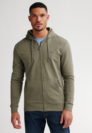 Sudadera verde oliva con cremallera, capucha ajustable, bolsillos frontales, puños acanalados y un pequeño logo en el pecho. Llevada sobre una camiseta azul.