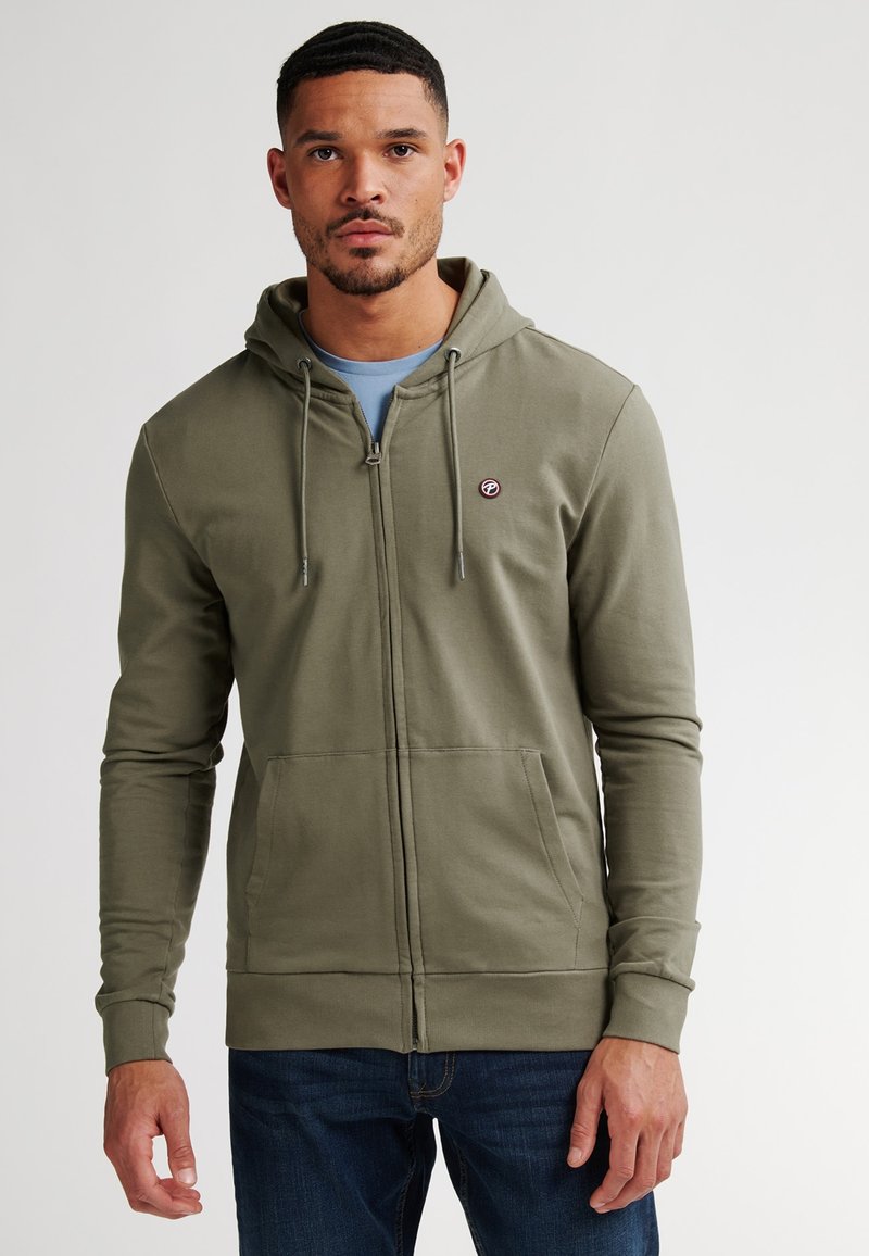 Felpa con zip di colore verde oliva, con cappuccio regolabile, tasche frontali, polsini a coste e un piccolo logo sul petto. Indossata sopra una maglietta blu.