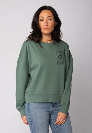Groene oversized sweatshirt met een ronde hals, voorzien van een zwarte zonafbeelding op de linkerborst en geribbelde manchetten en zoom. Dragers in combinatie met blauwe jeans.