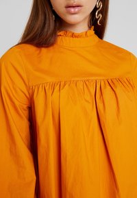 Oranssi paita, jossa on röyhelökaulus, kootut hihat ja pitkät hihat. Kankaassa on sileä pinta ja rento istuvuus.