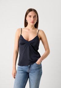 Marineblå satinkjole med tynne stropper, V-hals, rynkedetalj ved brystet og en glatt tekstur, parret med lyseblå jeans.
