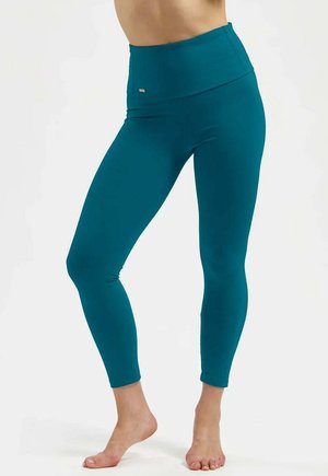 Femme portant des leggings taille haute couleur sarcelle, debout pieds nus avec les jambes légèrement croisées sur un fond uni.