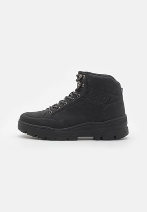 Botas negras de ante hasta el tobillo. Presentan ojales de metal, cordones y una suela de goma texturizada con un talón reforzado.