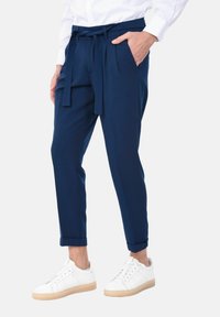 Pantaloni sartoriali blu navy con vita alta, pieghe frontali, cintura legata e tasche laterali, abbinati a sneaker bianche con suola color cuoio.