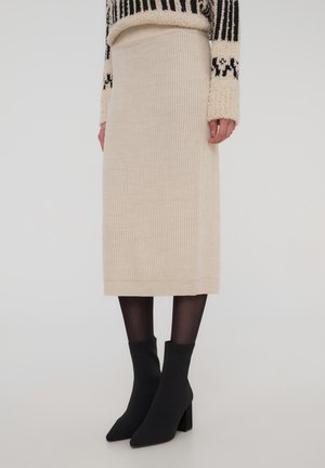 Gonna beige in maglia a coste con silhouette dritta, abbinata a stivaletti neri alla caviglia e collant neri trasparenti. Il tessuto ha una texture morbida.