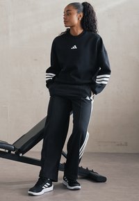 Sweatshirt de sport noir avec logo Adidas blanc, associé à un pantalon noir assorti avec des accents de bandes blanches. Le tout sur un fond neutre.