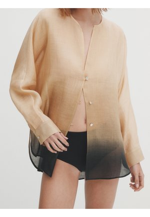 Kobieta ubrana w luźną, beżową koszulę z długim rękawem, która przechodzi w czerń na dole, rozpiętą, w połączeniu z czarnymi szortami, na jasnym tle.