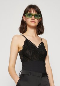 Camisole en dentelle noire avec bretelles spaghetti réglables, dotée d'une couche noire ajustée et de détails en dentelle complexes. Modèle portant des lunettes de soleil vertes.