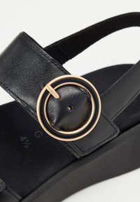 Gros plan sur une bride de sandale en cuir noir avec une boucle ronde noire et dorée sur une semelle compensée noire.