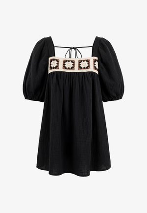 Zwarte jurk met pofmouwen en een vierkante halslijn, met een gehaakt bloemenband in crème, bruin en zwart over de borst en een detail met een strik aan de achterkant.