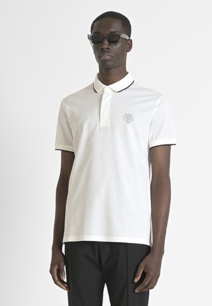 Polo bianco con colletto e bordi delle maniche neri, caratterizzato da un design ricamato sul petto. Materiale di cotone liscio.