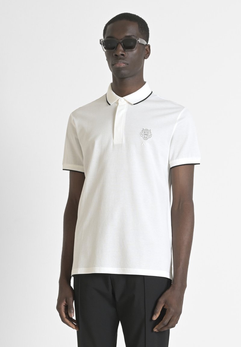 Camisa polo blanca con un cuello negro y ribetes en las mangas, que presenta un diseño bordado en el pecho. Material de algodón suave.