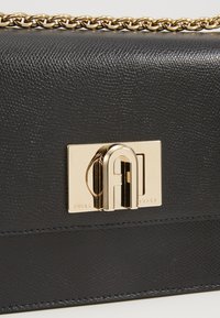 Borsa a mano in pelle nera con una finitura leggermente texturizzata, dotata di chiusura in metallo dorato e tracolla a catena. Dettaglio del logo evidente sulla parte anteriore.