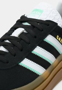 Zapatilla negra Adidas Gazelle con rayas blancas y verde menta, cordones negros, suela de goma y logo de Adidas en la lengüeta en verde menta.