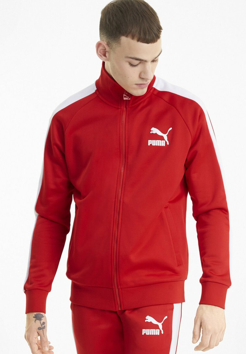 Veste de sport rouge en tissu lisse, dotée d'un col montant, d'une fermeture éclair intégrale, de panneaux latéraux blancs et d'un logo Puma sur la poitrine et la manche.