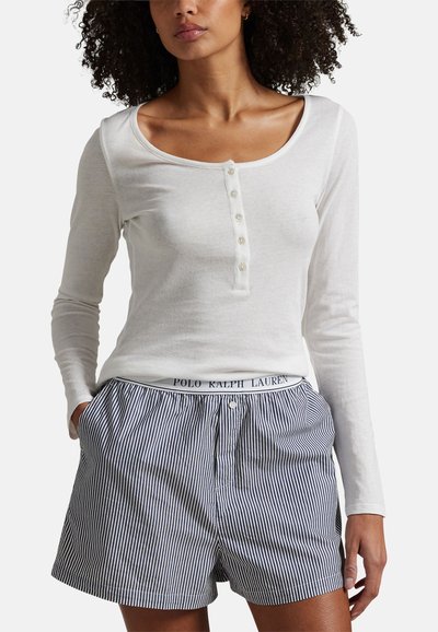 Vrouw die een wit, lang mouwig, knoopjeshemd draagt met blauw-wit gestreepte korte broek, handen in de zakken, torso zichtbaar van kin tot dijen.