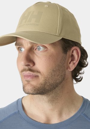 Hombre con barba clara mirando hacia la izquierda, con gorra beige de Helly Hansen y camiseta azul de cuello redondo contra un fondo liso.