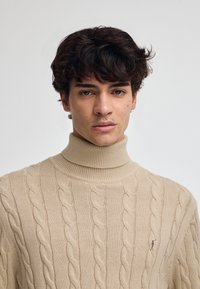 Beige polotröja med kabelstickat mönster, ribbad krage och mudd, med en liten broderad logotyp på bröstet.