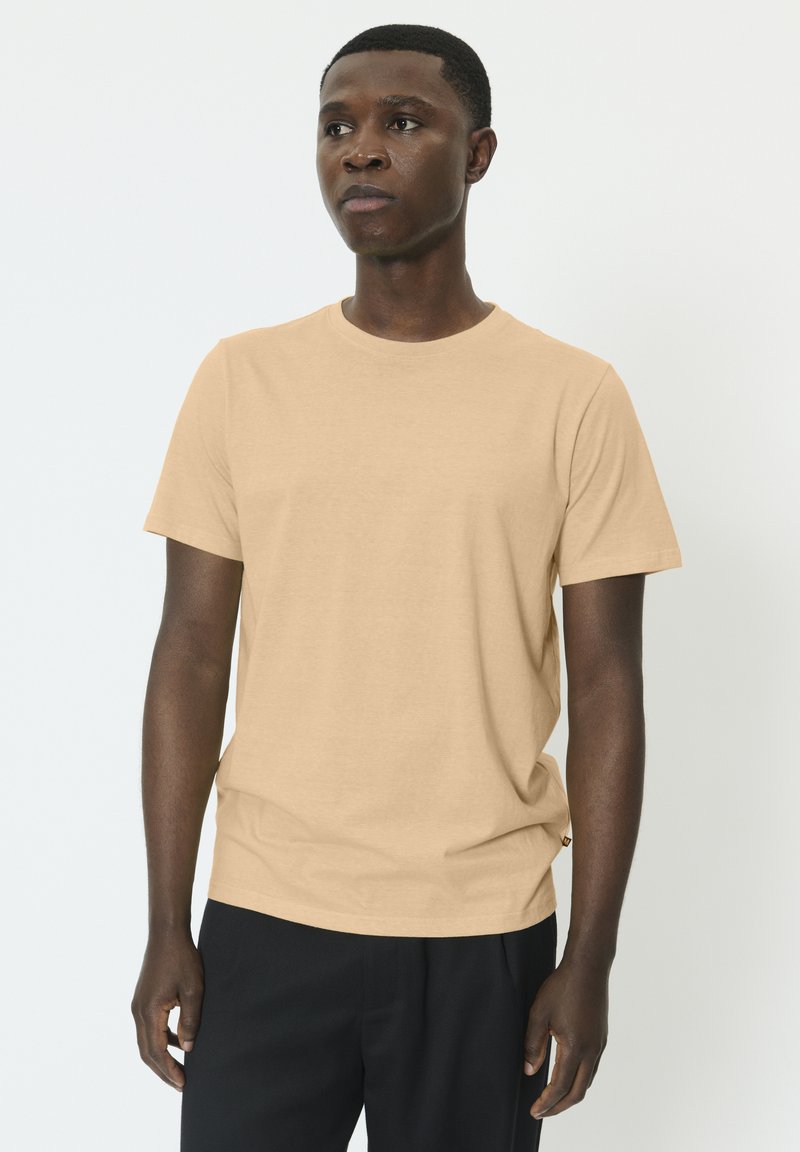 T-shirt beige à manches courtes en tissu doux et lisse avec un col rond. À noter un petit détail de logo à l'ourlet. Associé à un pantalon foncé.
