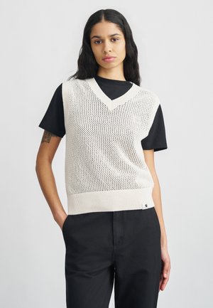 Gilet senza maniche a scollo a V lavorato a maglia color crema con una texture a rete, indossato sopra una t-shirt nera a maniche corte; abbinato a pantaloni neri.