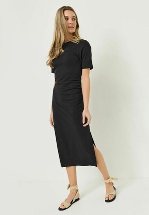 Žena s dlouhými blond vlasy, která má na sobě černé přiléhavé midi šaty s krátkými rukávy a béžové sandály s vázáním kolem kotníku, stojící před bílým pozadím.