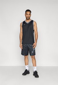 Čierna bezrukávová basketbalová dres s bielym lemovaním a logom, v kombinácii s čiernymi šortkami s cik-cak vzorom a červenými detailmi. Čierne tenisky.