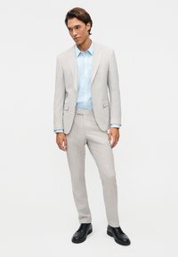 Jeune homme portant un costume gris clair avec un pantalon assorti, une chemise habillée bleu clair et des chaussures habillées noires, debout devant un fond blanc.