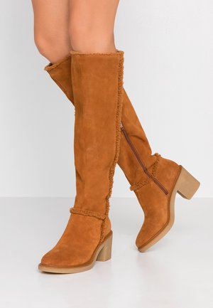 Kniehohe braune Wildlederstiefel mit Lammfellbesatz, Blockabsätzen und seitlichen Reißverschlüssen, getragen an nackten Beinen vor schlichtem Hintergrund.
