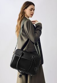 Sort læder skuldertaske med dobbelte håndtag og aftagelig rem. Udstyret med guldhardware, et diskret logo og en mønstret rem i grøn og sort.