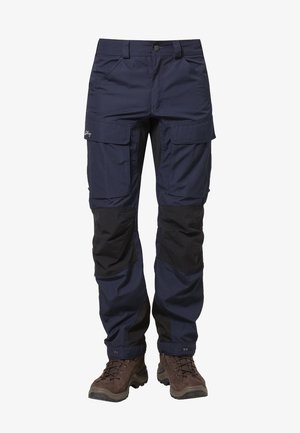 Pantalon cargo bleu marine avec renforts noirs aux genoux, poches avant à rabat, fermeture à boutons, porté avec des chaussures de randonnée marron.