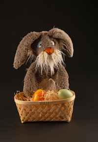 Lapin en peluche marron avec des oreilles tombantes et une barbe blanche, assis derrière un panier tressé contenant deux œufs de Pâques colorés sur de la paille.