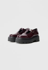 Zapatos Mary Jane de plataforma chunky en cuero burdeos oscuro brillante, con una correa de hebilla plateada y suelas gruesas de goma negra.