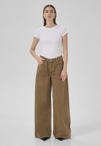 Camiseta blanca ajustada combinada con pantalones anchos marrones. La camiseta tiene mangas cortas y un sutil detalle bordado; los pantalones tienen un corte relajado y cordón.