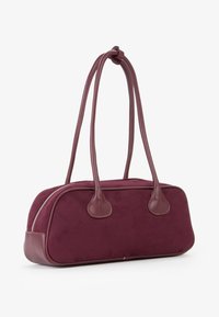 Borsa in suede bordeaux con dettagli in pelle liscia, chiusura a zip e manici doppi. Forma arrotondata, texture morbida e design minimalista.