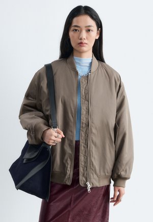 Femme aux longs cheveux foncés porte une veste bomber marron, un haut bleu et une jupe bordeaux, tenant un grand sac à bandoulière noir.