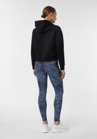 Czarna krótka bluza z kapturem i sznurkiem, zestawiona z legginsami w niebieski wzór leopardzi, o gładkiej fakturze. Na nogach białe buty sportowe.