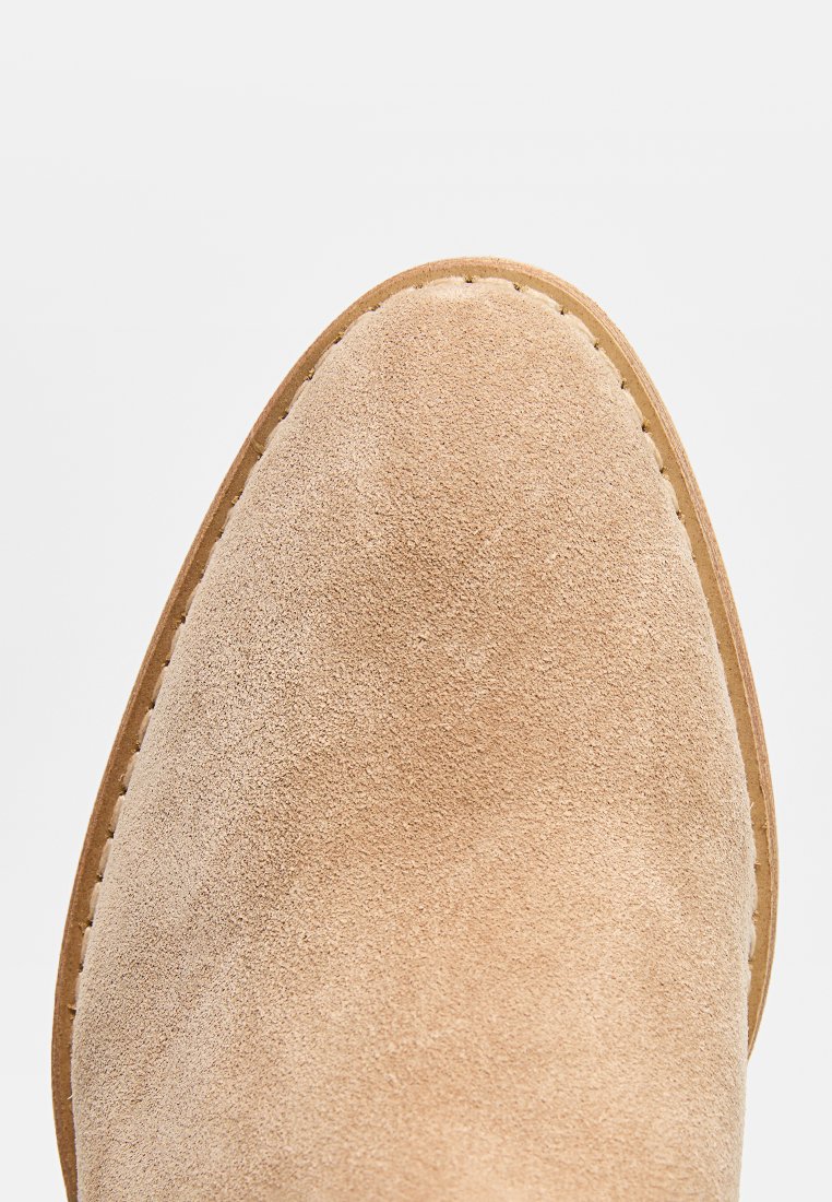 Zapato de ante beige claro con puntera redondeada, textura suave y suela de cuero marrón cosida. Sin herrajes ni patrones visibles.