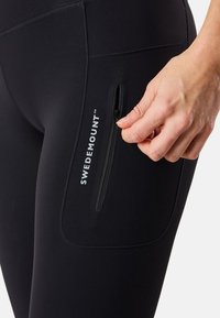 Leggings noirs avec une texture lisse, présentant une poche zippée et le logo "SWEDEMOUNT". Lignes épurées et design ajusté.