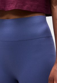 Άτομο που φοράει μπλε κολάν με υφή και ψηλή μέση και ένα κοντό μακρυμάνικο μπορντό τοπ, με το χέρι ακουμπισμένο στο πλάι.