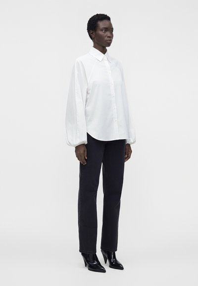 Femme debout portant une chemise blanche à manches longues boutonnée, un pantalon droit noir et des bottes noires à talons pointus sur un fond uni.
