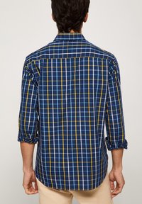 Košile s námořnickými modrými kostkami, které mají bílé a žluté mřížky, dlouhé rukávy vyhrnuté, a klasický límec. Textura bavlněné tkaniny.