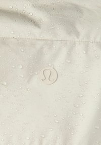 Wodoodporny beżowy materiał z kroplami, z małym, wypukłym białym logo. Powierzchnia gładka z delikatnym połyskiem.