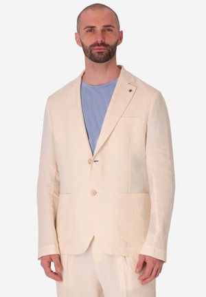 Uomo con la barba che indossa una giacca da completo color crema sopra una camicia azzurra, in piedi davanti a uno sfondo bianco semplice.