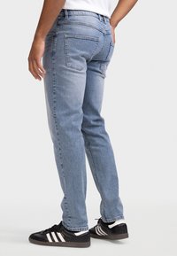 Φωτεινό μπλε τζιν με ίσια γραμμή, ορατές πίσω τσέπες και ξεθωριασμένη υφή. Συνδυασμένο με μαύρα αθλητικά παπούτσια με λευκές λεπτομέρειες.