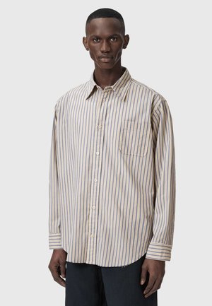 Männliches Modell trägt ein locker sitzendes Hemd mit vertikalen beigen, blauen und weißen Streifen, steht vor einem einfarbigen hellgrauen Hintergrund.