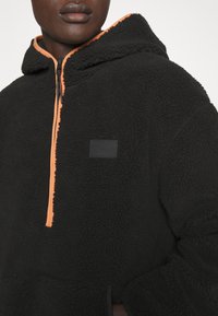 Pullover in pile nero con zip arancione, dotato di tasca a marsupio e patch logo nero sul petto. Tessuto testurizzato e morbido.