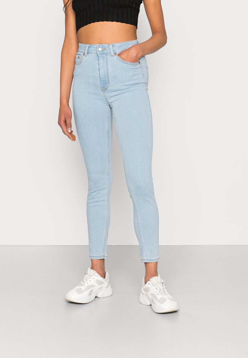 Jeans de cintura alta azul claro feitos de denim, design slim-fit com bolsos frontais, combinados com ténis brancos e uma blusa preta cropped.