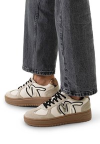 Sneaker mit oberem Teil in Creme, schwarzen und metallischen Akzenten, strukturiertem beigen Besatz, runder Zehenpartie und dicker beiger Laufsohle, kombiniert mit grauen Jeans.