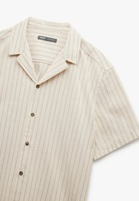 Camisa de manga corta en beige claro con rayas verticales marrón claro. Presenta un cuello y cinco botones marrón en la parte delantera. Tejido texturizado.